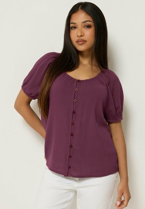 Femme aux longs cheveux foncés portant un chemisier violet à manches courtes boutonné et un pantalon blanc, debout devant un fond clair uni.