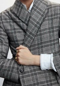 Grijze en zwarte geruite blazer met een dubbelzijdig ontwerp, met opgestikte revers en een gestructureerde stof. Witte shirtmanchet zichtbaar eronder.