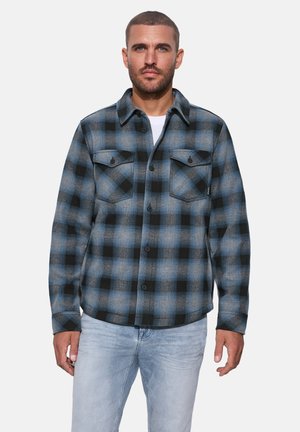Homme portant une chemise à carreaux bleue et noire boutonnée par-dessus un haut blanc et un jean bleu clair, faisant face avec une expression neutre.