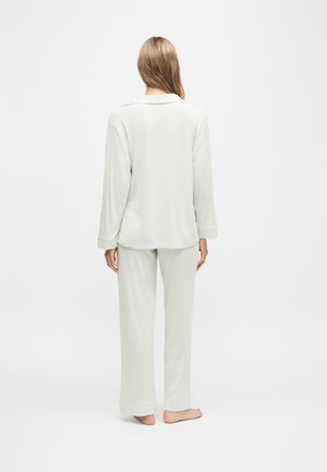 Femme aux longs cheveux blonds, debout pieds nus, de dos, portant un haut blanc ample à manches longues et un pantalon blanc assorti sur un fond uni.