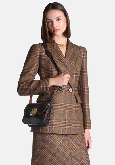 Femme portant un blazer à carreaux marron et une jupe, tenant un sac à épaule noir texturé avec une fermeture dorée sur fond blanc.