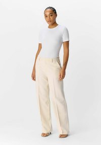 Haut blanc ajusté à manches courtes associé à un pantalon large beige taille haute et des sandales neutres, sur fond uni.