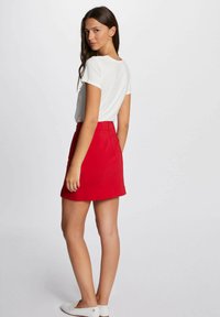 Gonna A-line rossa con chiusura a zip, abbinata a una maglietta bianca a maniche corte. Tessuto liscio e design pulito, indossata con scarpe bianche slip-on.