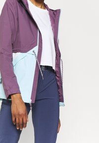 Chaqueta cortavientos en púrpura y azul claro, con cierre de cremallera, cordones ajustables y un diseño corto con puños elásticos.