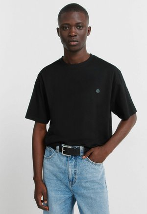 Jonge man die een zwart T-shirt met een klein logo en lichtblauwe spijkerbroek draagt, staande met één hand in zijn zak tegen een effen achtergrond.