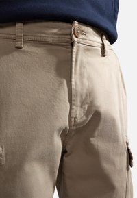 Lichtbeige katoenen broek met een gladde textuur, voorzien van een knoopsluiting, zijzakken en zichtbare stikdetails.