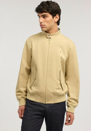 Giovane uomo che indossa una giacca beige con zip, colletto con bottoni a pressione e tasche con pattina, abbinata a jeans scuri, che guarda di lato.