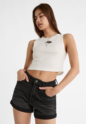 Jeune femme portant un débardeur blanc court avec un petit motif de chapeau et un short en jean noir taille haute, les mains dans les poches.