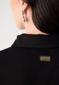 Zwart ribstof met een kraag, voorzien van een gouden en zwarte Barbour International-tag aan de achterkant. Soepele textuur en aansluitend ontwerp.