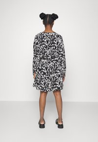 Schwarz-weiß gemustertes Kleid mit langen Ärmeln, tailliertem Bund und ausgestelltem Rock. Der Stoff wirkt glatt. Model trägt schwarze Schuhe.
