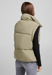 Kaki prošívaná vesta s vysokým límcem bez rukávů, s prošitým polstrováním a matným povrchem, nošená přes černý žebrovaný top.