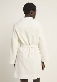 White textured bathrobe with a wrap design, wide sleeves, and a belted waist. Features a subtle horizontal pattern and a mid-thigh length.