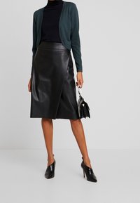 Woman wearing black leather knee-length skirt, black high heels, dark green cardigan, and holding a small black handbag.