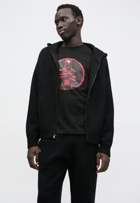 Czarna bluza z kapturem na zamek, na czarnej koszulce z czerwonym motywem Deadpoola, w połączeniu z czarnymi spodniami. Gładka tkanina i luźny krój.