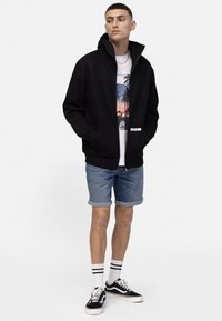Sweat à capuche noir zippé avec un col haut, associé à un t-shirt graphique, un short en denim et des baskets noires avec des accents blancs.