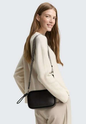 Sac en cuir noir en bandoulière de forme rectangulaire, avec une sangle tressée et une finition texturée, porté sur un pull de couleur crème.