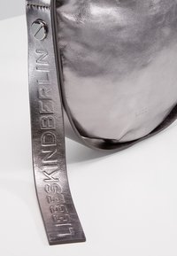 Metallinen hopeinen olkalaukku, jossa on sileä pinta, varustettuna kohokuvioidulla hihnalla ja pienellä hopeisella renkaalla.