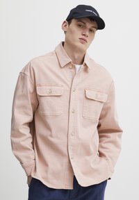 Világos rózsaszín farmering, két elülső zsebbel, gombos pánttal és hegyes gallérral. A modellen sötét sapka van, amelyen a "Casual Apparel" felirat olvasható.
