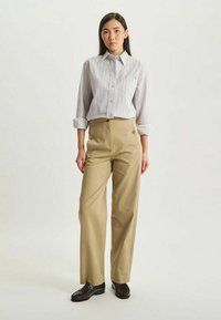 Chemise à rayures grise clair avec logo bleu ; pantalon taille haute beige avec boutons sur le côté ; chaussures marron ; debout sur un fond neutre.