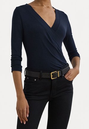 Vrouw die een nauwsluitend, marineblauw overslagshirt met lange mouwen en een diepe V-hals draagt, ingestopt in een zwarte broek en een zwarte riem met een gouden gesp.