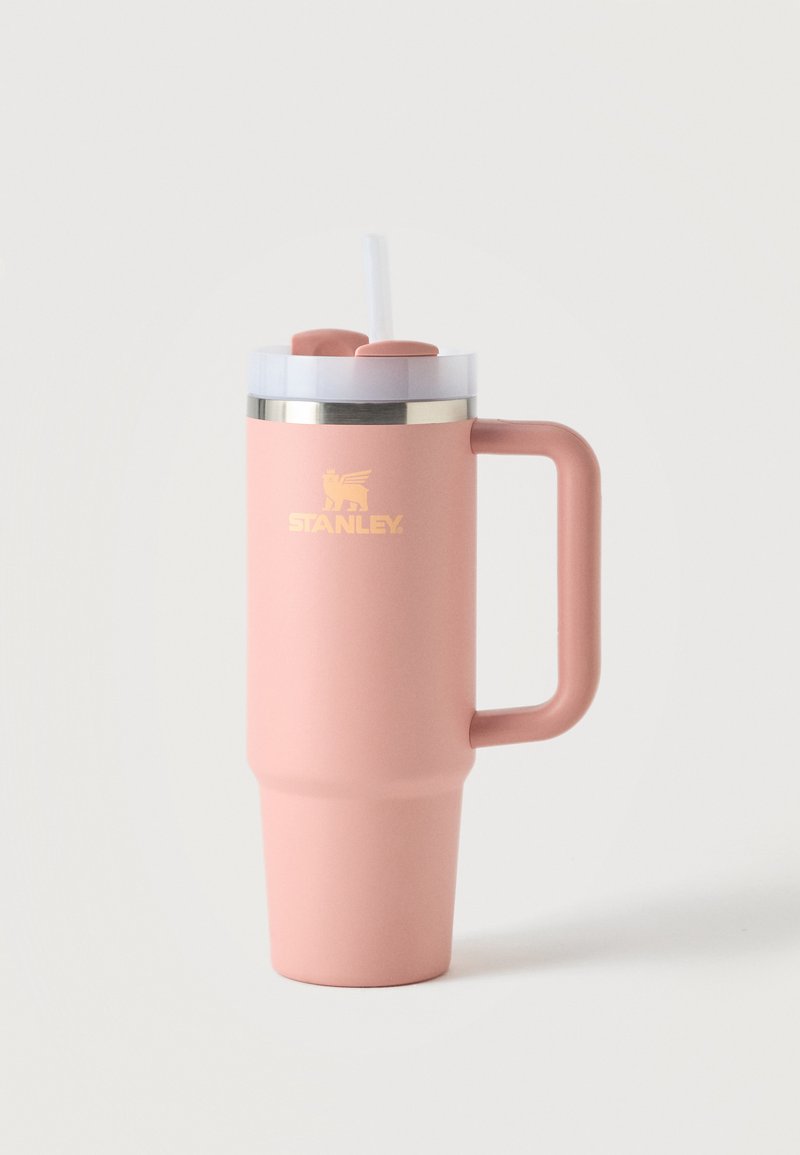 Cana de călătorie din oțel roz mat cu mâner, capac transparent, pai și logo Stanley auriu pe față, pe un fundal simplu și deschis.