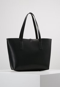 Sac cabas en cuir noir avec deux longues anses, une ouverture supérieure et une texture lisse. Présente un design minimaliste sans ornements supplémentaires.