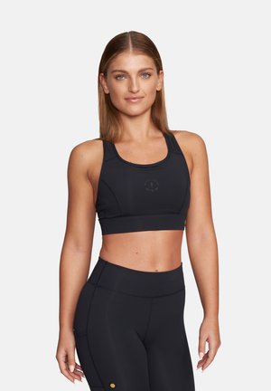 Junge Frau mit glattem Haar, die einen schwarzen Sport-BH und hoch taillierte schwarze Leggings trägt, steht vor einem schlichten Hintergrund.