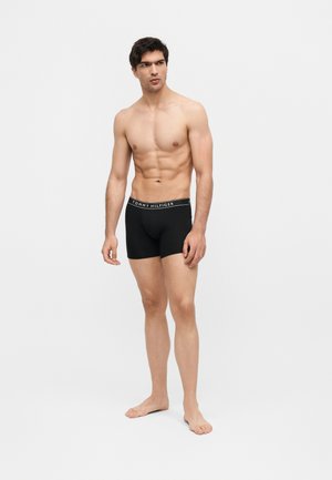 Άνδρας χωρίς πουκάμισο, που φοράει μαύρα boxer Tommy Hilfiger, όρθιος ξυπόλυτος σε απλό ανοιχτό γκρι φόντο, κοιτάζοντας αριστερά.