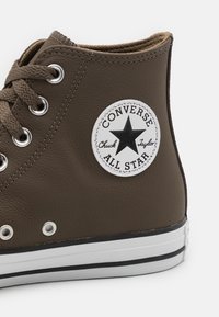 Sneaker alta marrone in pelle, con una toppa rotonda con logo nero e bianco, occhielli in metallo e suola in gomma bianca con accenti neri.