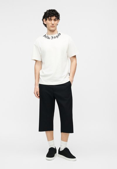 Νεαρός άνδρας στέκεται με το ένα χέρι στην τσέπη, φορώντας λευκό μπλουζάκι με μαύρα γράμματα στον γιακά, μαύρο κοντό παντελόνι, λευκές κάλτσες και μαύρα αθλητικά παπούτσια.