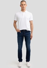 Muž stojící v bílé polokošili s krátkým rukávem, tmavě modrých úzkých džínách a bílých teniskách na jednobarevném pozadí.
