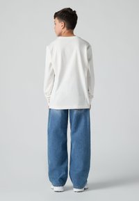 Λευκό μπλουζάκι με μακρύ μανίκι και κλειστό λαιμό, συνδυασμένο με φαρδιά μπλε τζιν παντελόνια. Τα τζιν έχουν σχέδιο διπλής τόνης στην τσέπη και κάθονται στη μέση.