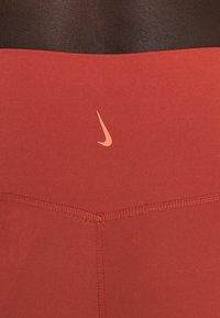 Punainen Nike korkeavyötäröinen legginsit, joissa on sileä pinta, tikatut saumat ja pieni beigekirjailtu Nike-logo vyötäröllä.