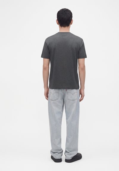 Homme debout de dos portant une chemise à manches courtes gris foncé, un jean ample bleu clair et des chaussures noires sur un fond blanc uni.