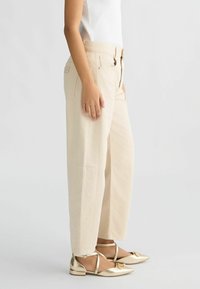 Pantalones de pierna ancha beiges hechos de algodón, con bolsillos delanteros y cintura alta, combinados con zapatos planos dorados con tiras y un acento decorativo.