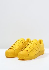 Zapatillas amarillas con un upper texturizado, una puntera lisa y tres rayas en el lateral. Diseñadas con cordones y una suela de goma.