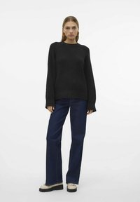 Pull en tricot noir avec texture côtelée, manches longues et col rond, associé à un jean évasé bleu foncé et des chaussures beiges.
