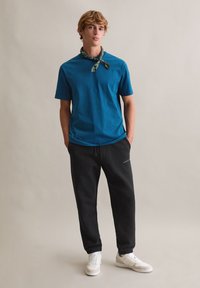 Turkusowy bawełniany t-shirt z krótkimi rękawami, zestawiony z czarnymi spodniami dresowymi z akcentem logo oraz białymi trampkami. Widoczna tekstura tkaniny.