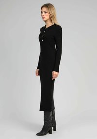 Robe noire en tricot côtelé avec col polo et patte de boutonnage à cinq boutons. Silhouette ajustée avec des manches longues et un ourlet légèrement évasé.