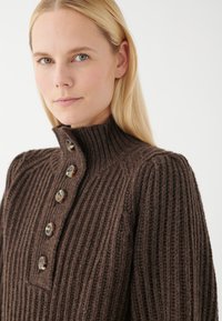 Brauner gerippter Strickpullover mit hohem Kragen und fünf Knopfverschluss, ausgestattet mit strukturiertem Stoff und subtilen Farbvariationen.