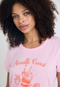 T-shirt de algodão rosa com as palavras "Costa de Amalfi" em texto laranja, acompanhada de uma ilustração de um cocktail e detalhes de frutas. Mangas curtas e decote redondo.