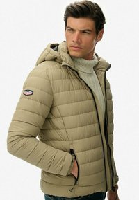 Giacca puffer khaki con cappuccio, chiusura con zip, design a righe verticali e tasche laterali. Presenta una patch con il logo sulla manica sinistra.