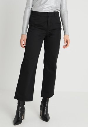 Femme portant un pantalon noir à jambes larges, un haut argenté à manches longues et des bottines noires à talons, debout devant un fond uni.