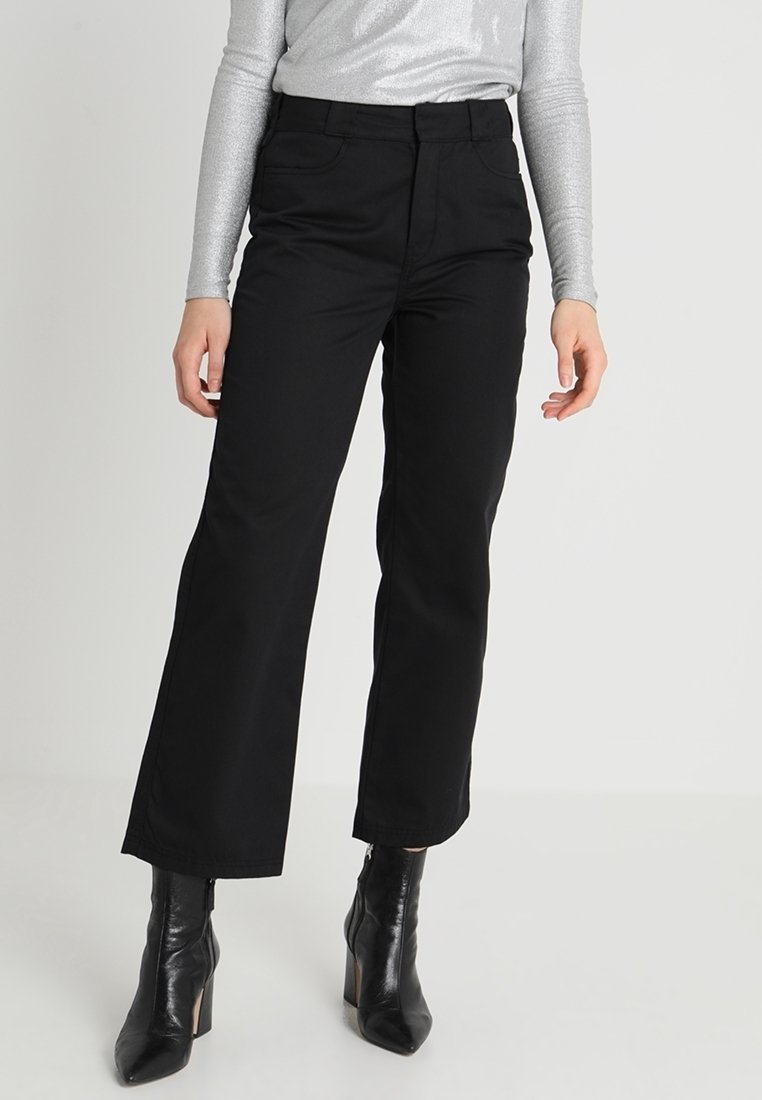 Femme portant un pantalon noir à jambes larges, un haut argenté à manches longues et des bottines noires à talons, debout devant un fond uni.