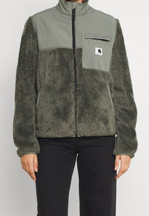 Personne portant une veste polaire verte zippée avec tissu texturé, poche poitrine avec logo Carhartt et pantalon noir.
