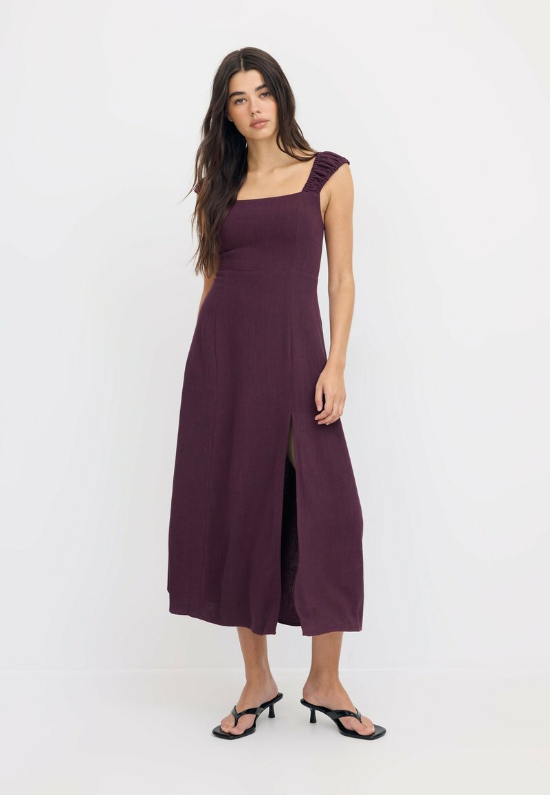 Femme portant une robe midi sans manches violet foncé avec des bretelles froncées et une fente sur le devant, associée à des sandales noires à talons.