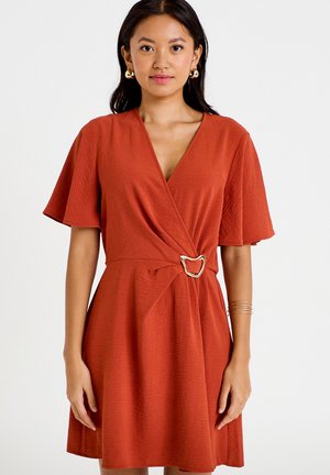 Femme portant une robe portefeuille orange rouille à manches courtes avec une ceinture à boucle dorée, accessoirisée de boucles d’oreilles créoles et de bracelets dorés.