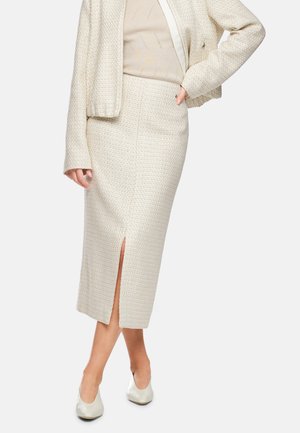 Woman wearing a cream textured midi skirt with front slit, matching jacket, beige top, and white low-heeled shoes standing with one hand on hip.