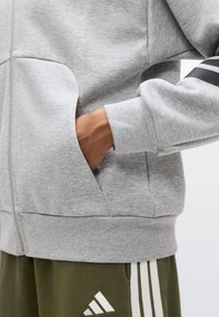 Γκρι φούτερ με φερμουάρ, πλαϊνές τσέπες, ριμπ μανσέτες και υφή τελειώματος. Διαθέτει μαύρες ριγέ λεπτομέρειες στα μανίκια και άνετη εφαρμογή.