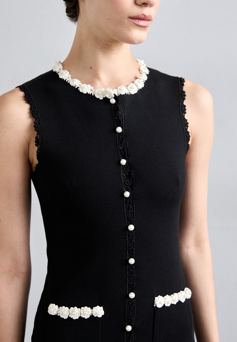 Schwarzes Kleid mit einem Kragen aus floraler Spitze, Perlenknöpfen und weißen Blumenakzenten an Taschen und Schultern. Ärmelloses Design mit strukturierter Stoff.